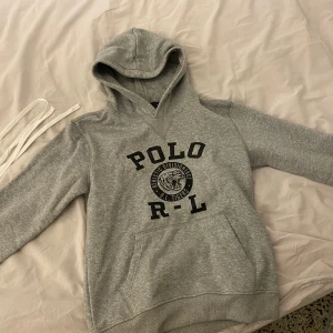 Grå hoodie från Polo Ralph Lauren - Säljer en grå hoodie från Polo Ralph Lauren med ett sjukt snyggt tryck på magen, väldigt stilren och snygg hoodie. Vill egentligen behålla men säljer pga att den är för liten. Inga defekter eller liknande och mycket sällan använd. Tveka inte på att ställa frågor😄