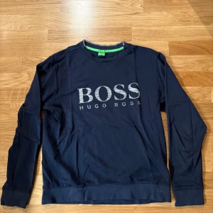 Marinblå Hugo Boss sweatshirt - Snygg marinblå sweatshirt från Hugo Boss med stort BOSS-tryck i silver på bröstet. Tröjan har lång ärm, ribbade muddar och rund hals. Slim fit-modell som sitter schysst på kroppen. Perfekt för dig som gillar stilrena märkesplagg.