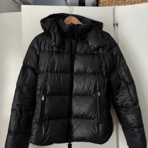 Svart dunjacka från BOSS med huva - Svart pufferjacka från BOSS med äkta dunfyllning och stor, mysig huva. Jackan har två sidofickor med dragkedja och en clean, quiltad look. Perfekt för kalla dagar och har en snygg, modern passform. Stängs med dragkedja framtill.