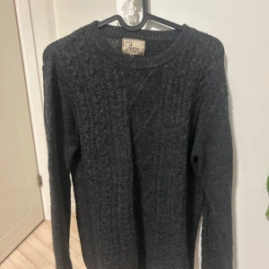 Mörkgrå stickad tröja från Ashley - Mysig mörkgrå stickad tröja från Ashley med klassiskt kabelstickat mönster. Rund halsringning och långa ärmar gör den perfekt för kyliga dagar. Passar dig som gillar enkel och stilren design med lite extra struktur.