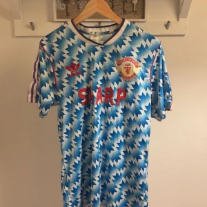 Manchester United retro bortatröja Adidas - Retro Manchester United bortatröja från Adidas med ikoniskt blått och vitt blixtmönster, röda detaljer och klubbmärke på bröstet. Klassiska Adidas-ränder på axlarna och SHARP-logga framtill. Tillverkad i lätt och ventilerande material, perfekt för fotbollsfans.