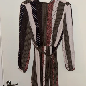 Randig långklänning från Mohito - Säljer en långklänning från Mohito med snygga vertikala ränder i brunt, rosa, beige och svart. Klänningen har lång ärm, v-ringning och ett matchande bälte i midjan. Materialet är mjukt och flowy, perfekt för dig som gillar mönster och en relaxed men chic look.