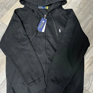 Svart hoodie från Polo Ralph Lauren - Svart hoodie från Polo Ralph Lauren med klassisk logga broderad i vitt på bröstet. Tröjan har huva med snörning, dragkedja framtill och ribbade muddar. Tillverkad i mjuk bomull för skön komfort. Perfekt för en chill och stilren look.