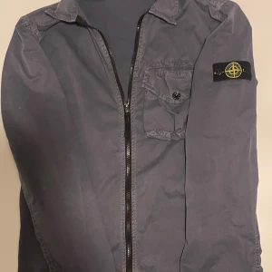 Blå overshirt från Stone Island - Säljer emot min vilja en blå overshirt från Stone Island med klassisk logotyp på vänster ärm. Jag säljer nu denna då den börjar bli för liten. Jackan har en bröstficka med knapp, dragkedja framtill och krage. Materialet är lätt och syntetiskt, perfekt för lager-på-lager. Passformen är normal och ärmarna är långärmade. Jackan passar för er som har storlek XS/S. Priset är ej hugget i sten! Ställ om det vore några frågor!