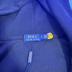 Svart hoodie från Polo Ralph Lauren - Svart hoodie från Polo Ralph Lauren med klassisk röd logga på bröstet. Tröjan har huva med snörning, dragkedja framtill och två fickor. Materialet är mjuk bomull och passformen är normal. Perfekt för chill dagar och streetwear vibes.