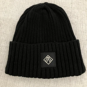 Svart ribbad mössa från Marengo - Svart ribbstickad mössa från Marengo Sweden med uppvikt kant och stilren logotyp framtill. Klassisk form som passar perfekt till kalla dagar. Tillverkad i mjuk akryl för skön komfort.