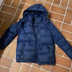 Mörkblå pufferjacka från Polo Ralph Lauren - Säljer en mörkblå pufferjacka från Polo Ralph Lauren med huva och dragkedja framtill. Jackan har quiltad design, två sidofickor med dragkedja och en liten röd logga på bröstet. Perfekt för kalla dagar och har en snygg, normal passform.