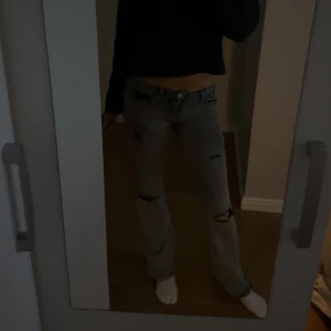 Grå slitna jeans med raka ben - Säljer ett par grå jeans med snygga slitningar och hål på benen. Modellen har låg midja och utsvängda💕