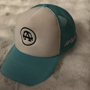Turkos och vit keps från All Cap One - Snygg truckerkeps från All Cap One med turkos mesh och vit frontpanel. Framsidan har ett broderat svart logomärke och sidan är broderad med 'All Cap One'. Skärmen är turkos och kepsen har justerbar passform. Perfekt för dig som vill sticka ut med en fräsch färg.
