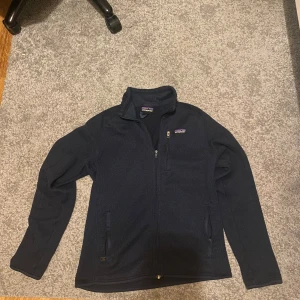 Svart fleecejacka från Patagonia - Mjuk och varm svart fleecejacka från Patagonia med hel dragkedja framtill, hög krage och tre fickor med dragkedja. Jackan har en stilren look och passar perfekt för kyliga dagar. Diskret Patagonia-logga på bröstet.