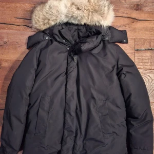 Svart dunjacka med päls från Canada Goose - Svart dunjacka från Canada Goose med stor fluffig päls på huvan och klassiska Arctic Program-märken på ärmen och insidan. Jackan har två stora fickor fram, dragkedja och knappar, samt en rejäl huva som skyddar mot kyla. Perfekt för kalla vinterdagar.