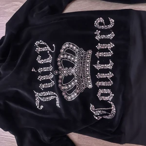 Svart velour hoodie Juicy Couture XS - Svart hoodie från Juicy Couture i mjuk velour med glittrande strassdetaljer och krona på ryggen. Dragkedja framtill, huva och långa ärmar. Perfekt för dig som gillar ikonisk Y2K-stil och vill sticka ut med bling.