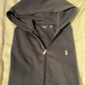 Svart hoodie från Polo Ralph Lauren - Svart hoodie från Polo Ralph Lauren med klassisk huva och dragkedja framtill. Tröjan har det ikoniska Polo-logot broderat i vitt på bröstet. Tillverkad i mjuk bomull för skön komfort och avslappnad stil. Perfekt för dig som gillar stilrena och enkla plagg.