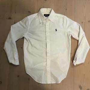 Vit skjorta från Polo Ralph Lauren - Klassisk vit skjorta från Polo Ralph Lauren i bomull med broderad marinblå logga på bröstet. Skjortan har lång ärm, knappar framtill och klassisk krage. Passformen är classic fit och materialet känns mjukt och skönt.