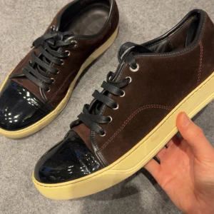 Lanvin sneakers i mocka och lack uk8 - Snygga sneakers från Lanvin i mörkbrun mocka med svarta lackdetaljer på tån och svarta skosnören. Skorna har en beige platt sula och rund tå. Perfekt för dig som vill ha en stilren men ändå unik sneaker med lyxig känsla.