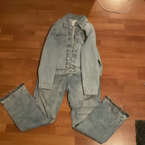 Ljusblå denimkostym med fransade ben - Säljer en ljusblå denimkostym med kavaj och matchande jeans. Jeansen har fransade benslut och en loose passform. Kavajen har klassisk krage och knappar framtill. Perfekt för dig som gillar retro och streetstyle.