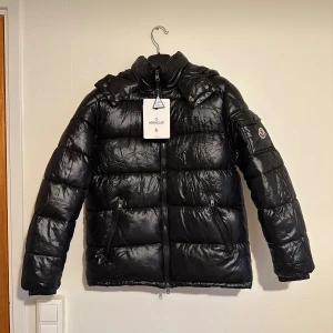 Svart pufferjacka från Moncler S - Snygg svart pufferjacka från Moncler med glansig finish och klassisk logga på ärmen. Jackan har dragkedja framtill, två fickor med dragkedja och hög krage. Insidan har ett coolt serietryck. Perfekt för dig som vill ha en stilren och varm vinterjacka Jackan är aldrig använd! Pris kan såklart diskuteras, tveka inte kolla mina omdömen ☺️