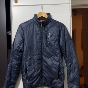 Marinblå pufferjacka från Sail Racing - Snygg marinblå pufferjacka från Sail Racing med hög krage, hel dragkedja och två fickor med orange detaljer. Jackan har Sail Racing-logga på bröstet och ett märke på ärmen. Perfekt för kyliga dagar och stilren look.