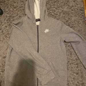 Grå Nike hoodie med dragkedja XL - Säljer en grå hoodie från Nike i storlek XL. Tröjan har huva, hel dragkedja framtill och två fickor. Klassisk Nike-logga i vitt på bröstet. Perfekt för chill eller träning, standard fit och mjukt material.