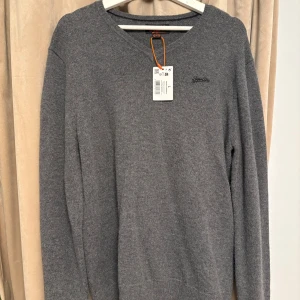 Grå stickad tröja från Superdry - Snygg grå stickad tröja från Superdry med diskret logga på bröstet. Tröjan har v-ringad hals och långa ärmar, perfekt för lager-på-lager. Mjuk och skön känsla, passar till jeans eller chinos.