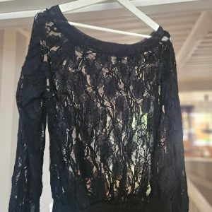 Svart spetsblus med lång ärm - Säljer en svart blus i transparent spets med blommigt mönster. Blusen har lång ärm och bred halsringning, perfekt för dig som gillar en elegant och edgy stil. Materialet är mjukt och följsamt, och spetsen ger en cool vibe.