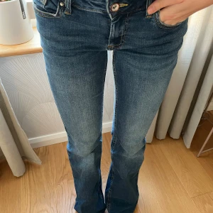 Bootcut jeans med låg midja - Säljer ett par klassiska blå jeans med bootcut-modell och låg midja. Jeansen har fem fickor, snygg tvätt och sitter tajt upptill med utsvängda ben. Midjemått ca 37cm o innerbenslängd ca 73cm, skulle säga att de motsvarar ca storlek 34/36