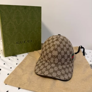 Gucci beige keps med GG-mönster - Snygg beige keps från Gucci med det klassiska GG-monogrammet över hela kepsen. Kepsen har en böjd skärm och justerbar rem baktill. Materialet är bomull och polyester, och på sidan finns Guccis ikoniska gröna och röda ränder. Perfekt för dig som vill ha en lyxig streetstyle. Äkta!!! Mer bilder finns privat! 