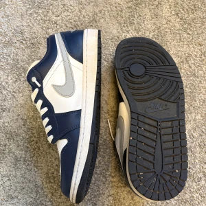 Nike Jordan 1 Low marinblå/vit 43 - Nike Jordan 1 Low sneakers i marinblått och vitt med silverfärgade detaljer. Klassisk rund tå, snörning och platt sula. Swoosh-logga i silver på sidan och Air Jordan-märke på hälen. Ovandel i skinn och syntet, perforerad tå för extra ventilation.