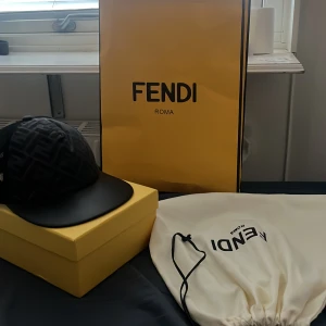 Svart Fendi keps med FF-mönster - Säljer en svart keps från Fendi med ikoniskt FF-mönster på framsidan. Kepsen har mesh-paneler bak för extra ventilation, justerbart spänne med Fendi-logga och böjd skärm i svart. Kommer med originalbox och dustbag. Perfekt för dig som vill sticka ut med lyxig streetstyle.