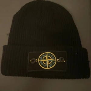 Svart ribbstickad mössa Stone Island - Svart ribbstickad mössa från Stone Island med klassisk uppvikt kant och den ikoniska logotypen framtill. Märket är broderat i gult och grönt på svart patch som sitter fast med två knappar. Perfekt accessoar för kalla dagar.