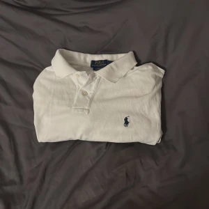 Vit pikétröja från Polo Ralph Lauren - Vit pikétröja från Polo Ralph Lauren med klassisk krage och två knappar framtill. Tröjan har det ikoniska blå broderade logotypen på bröstet och är tillverkad i mjuk bomull. Perfekt för en clean och stilren look. Jag har knappt använt den och varit väldigt försiktig med den. Köpte ny för 1499 på care of Carl 