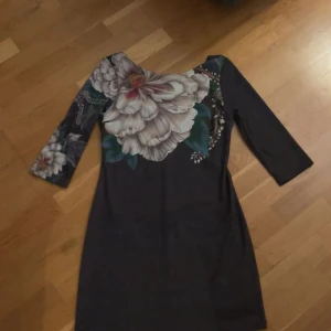 Mörk blommig Desigual klänning - Snygg mörk klänning från Desigual med stora blommor i vitt och beige på axel och bröst. Klänningen har trekvartsärm, rundad hals och en figurnära passform. Materialet är mjukt och stretchigt, perfekt för dig som gillar unika mönster och färgkombinationer.