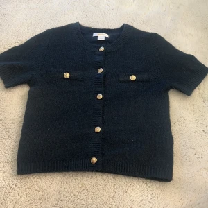 Marinblå stickad blus från Gina Tricot - Marinblå stickad blus från Gina Tricot med korta ärmar och rund hals. Framsidan har dekorativa guldknappar och två fejkade bröstfickor. Ribbstickade kanter ger en snygg finish. Perfekt för dig som gillar klassisk men trendig stil.