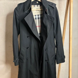 Burberry trenchcoat  - Säljer min äkta Burberry trenchcoat. På bilderna syns märkets originaletiketter som bekräftar att den är genuin. Kappan är i mycket gott skick utan synliga defekter på utsidan – inga lösa trådar, hål eller liknande. Jag bedömer att den har ett mycket välvårdat utseende och ser nästintill ny ut, även om den är använd. Det står i kappan att det är storlek 6 vilket jag förmodar är storlek XS-S. Kappan levereras med ett avtagbart foder som gör den varmare vid behov. Själva fodret har dock några mind