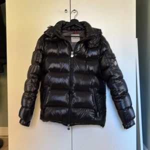 Svart pufferjacka herr - Säljer en svart, glansig pufferjacka med huva och dragkedja. Jackan har logga på ärmen, praktiska fickor och quiltad design. Perfekt för kalla dagar och riktigt snygg streetstil. Storlek L men passar M