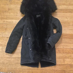 Svart parkasjacka med fuskpäls - Säljer en svart parkasjacka från Atelier med stor huva och mjukt fuskpälsfoder. Jackan har dragkedja och knappar framtill, samt elastiska muddar vid ärmsluten. Perfekt för kalla dagar med sin långa modell och fluffiga insida.