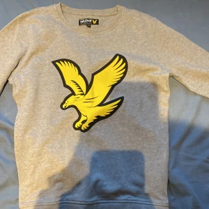 Grå sweatshirt från Lyle & Scott - Säljer en grå sweatshirt från Lyle & Scott med stort gult fågeltryck på bröstet. Tröjan har rund halsringning och långa ärmar. Tillverkad i mjuk bomull, perfekt för en chill och stilren look.