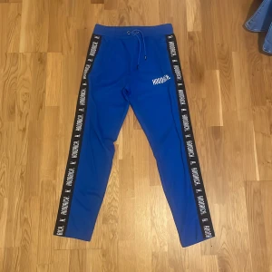 Blå mjukisbyxor från Hoodrich - Blå mjukisbyxor från Hoodrich med svarta sidor och vit logga på vänster ben. Byxorna har snörning i midjan och textdetaljer längs benen. Perfekta för en avslappnad och sportig stil.