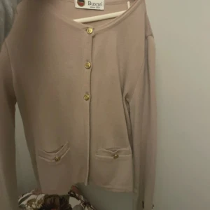 Dimrosa kofta från Busnel med guldknappar - Supersnygg rosa/beige kofta från Busnel med klassiska guldknappar framtill, på fickorna och vid ärmsluten. Koftan har två små fickor fram och rund halsringning. Ikonisk Busnel-logga på ena ärmen. Perfekt för lager-på-lager under höst och vår. Fint skick storlek 34
