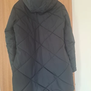 Svart quiltad lång pufferjacka - Säljer en svart lång pufferjacka med huva och dragkedja framtill. Jackan har ett quiltat mönster i stora rutor och två snedställda fickor med dragkedja. Perfekt för kalla dagar när du vill hålla dig varm och stilsäker.