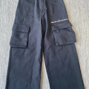Svarta cargobyxor från Miss S Denim - Svarta cargobyxor från Miss S Denim med breda ben och stora fickor på sidorna. Byxorna har dragkedjedetalj på ena fickan och är tillverkade i bomull. Perfekta för en avslappnad och trendig look. Mått Midjan: ca 38 cm Längd från grenen och ner: ca 71 cm  100% bomull  Kommer från ett djur och rökfritt hem. 