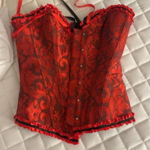 Röd och svart korsett med snörning - Säljer en snygg röd korsett med svarta detaljer och vackert mönster. Korsetten har svarta satinband för snörning i ryggen, knäppning fram och volangkant upptill och nedtill. Pris kan diskuteras 