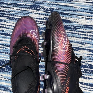 Puma Future Z lila fotbollsskor - Säljer ett par Puma Future Z fotbollsskor i svart och lila med orange detaljer. Skorna har snörning, stickad strumpkrage och en cool mönstrad ovandel. Sulan har lila dobbar för gräsplan. Perfekt för dig som vill sticka ut på planen!