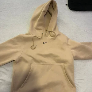Beige hoodie från Nike med huva - Säljer en beige hoodie från Nike med klassisk svart swoosh på bröstet. Tröjan har stor känguruficka, snörning i huvan och ribbade muddar vid ärmslut. Perfekt för chill dagar och streetwear-stil. Materialet är mjuk bomull och polyesterblandning. Funkar som bäst på vintern