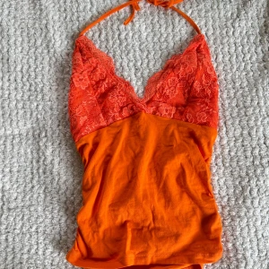 Orange spetslinnetopp med knyt - Snygg orange linnetopp med spetsdetaljer upptill och knyt runt nacken. Djup v-ringning och blommig spets ger en feminin vibe. Perfekt för dig som vill sticka ut med färg och detaljer.
