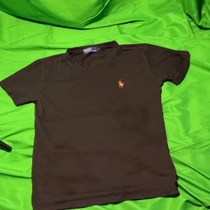 Svart t-shirt från Polo Ralph Lauren - Svart t-shirt från Polo Ralph Lauren med klassisk röd broderad logga på bröstet. T-shirten har rund hals och korta ärmar, tillverkad i mjuk bomull för en skön känsla. Enkel och stilren design som funkar till det mesta.Liknande modell som originalet!