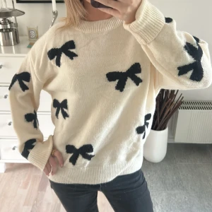 Beige stickad tröja med svarta rosetter - Superfin beige stickad tröja med svarta rosetter över hela. Tröjan har rund halsringning, ribbade muddar och lång ärm. Perfekt för dig som vill ha en söt och trendig look med en twist.