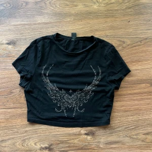 Svart crop top - Svart croppad topp från SHEIN i storlek XS. Toppen har korta ärmar och ett coolt mönster framtill gjort av silvriga nitar som bildar ett hjärtformat motiv med vingar. Perfekt för dig som gillar edgy stil och vill sticka ut. Några nitar har trillat av men förutom det så är den i ganska bra skick😁