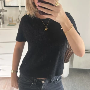 Svart topp med guldknappar från H&M - Svart kortärmad topp från H&M i storlek S, tillverkad i ett mjukt och lite fluffigt material. Toppen har rund hals och dekorativa guldknappar på axeln som ger en snygg detalj. Passformen är normal och toppen är enkel att matcha med jeans eller kjol.