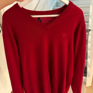 Röd v-ringad tröja från Gant - Röd v-ringad tröja från Gant med diskret broderad logga på bröstet. Tröjan är i mjuk bomull, har långa ärmar och ribbade muddar vid hals, ärmslut och nederkant. Perfekt för lager-på-lager och snygg till jeans eller kjol.